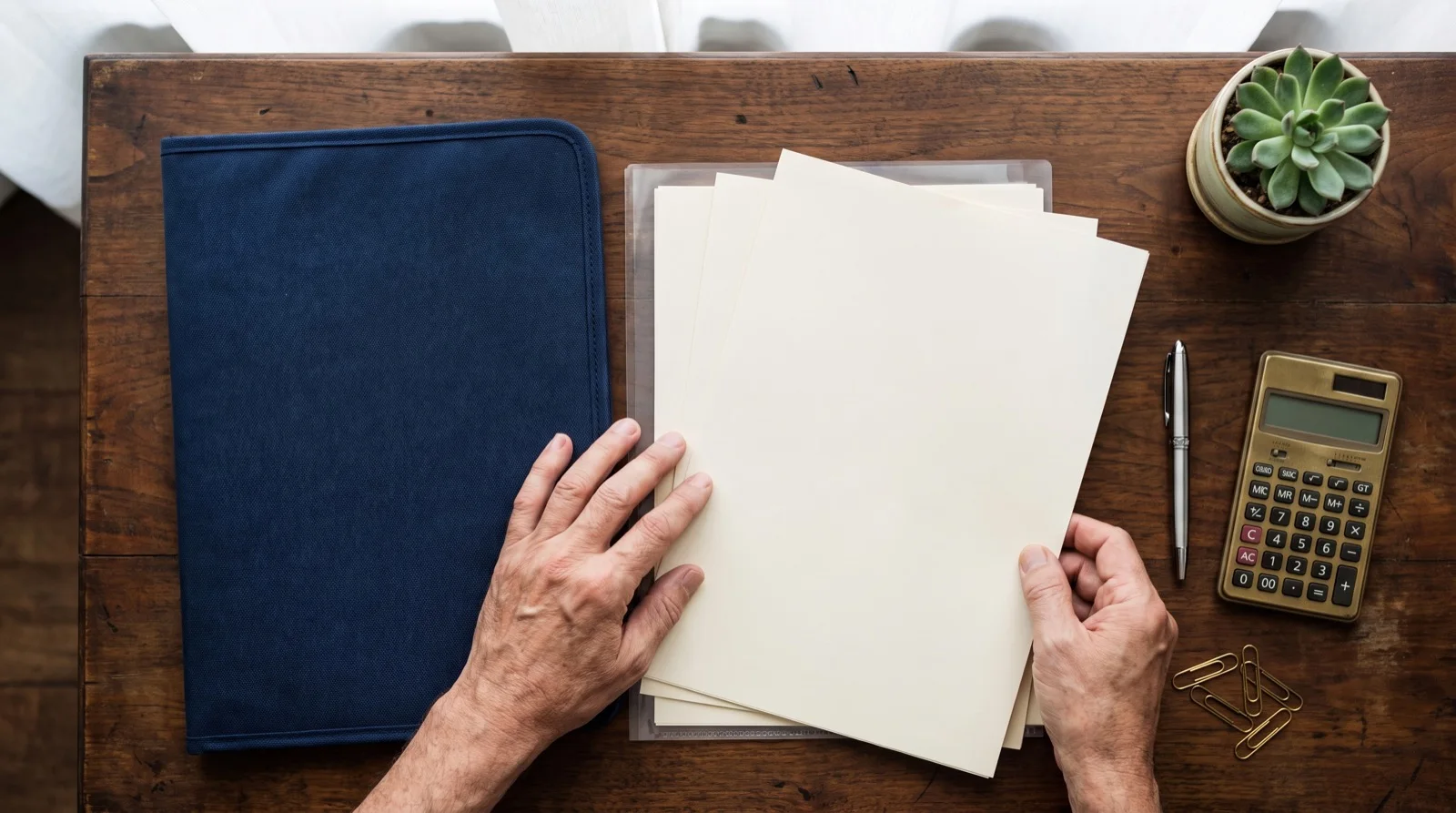 Hands organizing estate paperwork, a navy folder, a calculator and a pen on a wooden desk